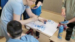 Na zdjęciu uczniowie malują dłonie niebieską farbą i odbijają ją na białym kartonie